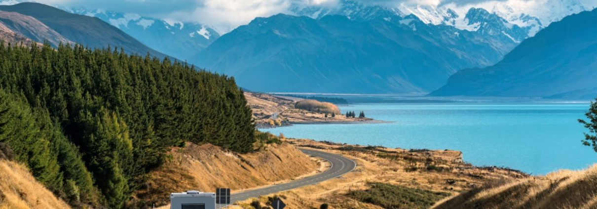 A motor home driving along a winding road beside a lake, with New Zealand's Southern Alps in the background