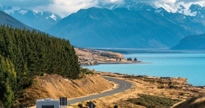 A motor home driving along a winding road beside a lake, with New Zealand's Southern Alps in the background