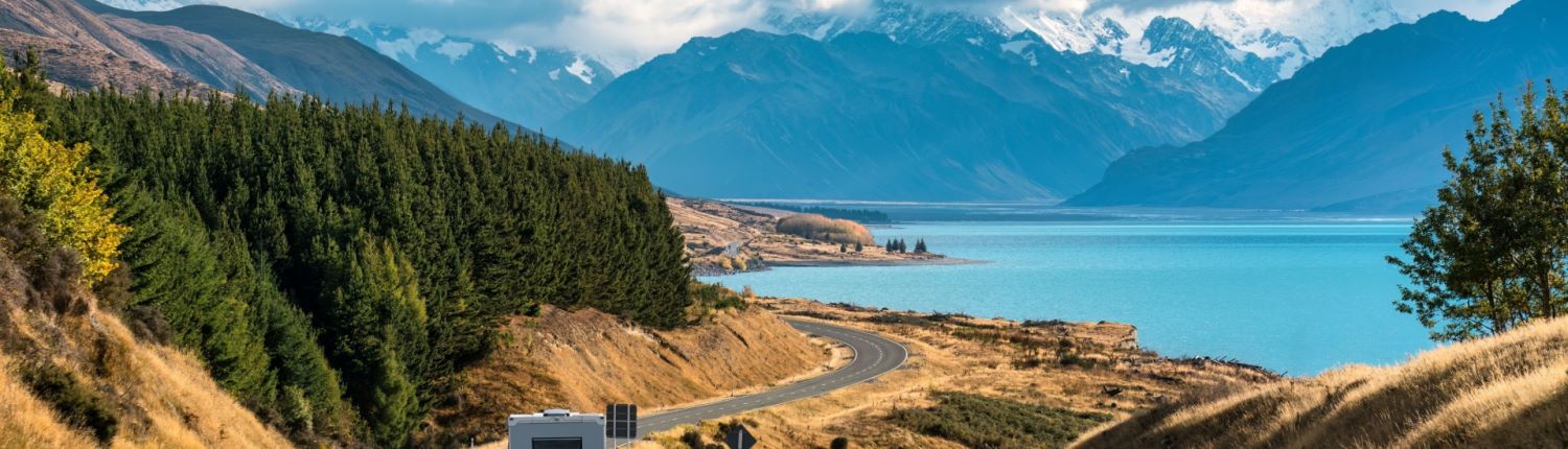 A motor home driving along a winding road beside a lake, with New Zealand's Southern Alps in the background