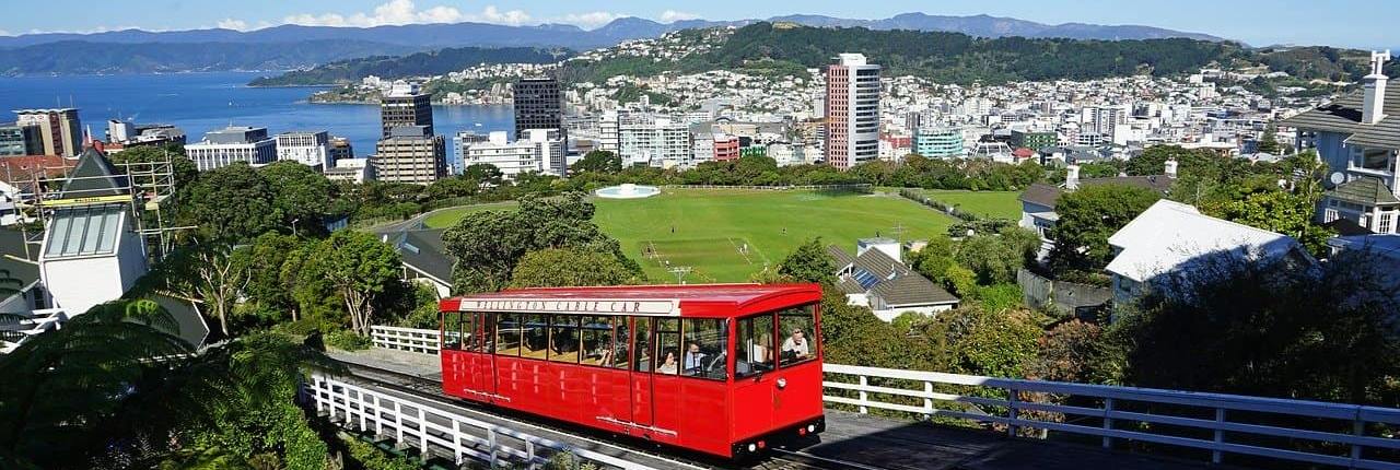 A red funicular railway car ascends a hill, overlooking a large cricket pitch and the city of Wellington, New Zealand