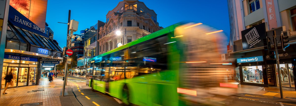 A blurred bus travels past shops on a street in Wellington, New Zealand