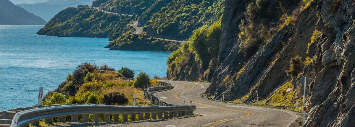 A winding road hugs a cliffside as it heads downhill towards a lakeside